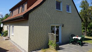 Une maison à deux étages, avec un toit en pente, une façade en briques et un petit porche à l’avant, agrémenté d’une table et de chaises.