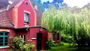 Ein rotes Backsteinhaus mit grüner Tür und Fenstern, umgeben von üppigem Grün und einer Trauerweide.