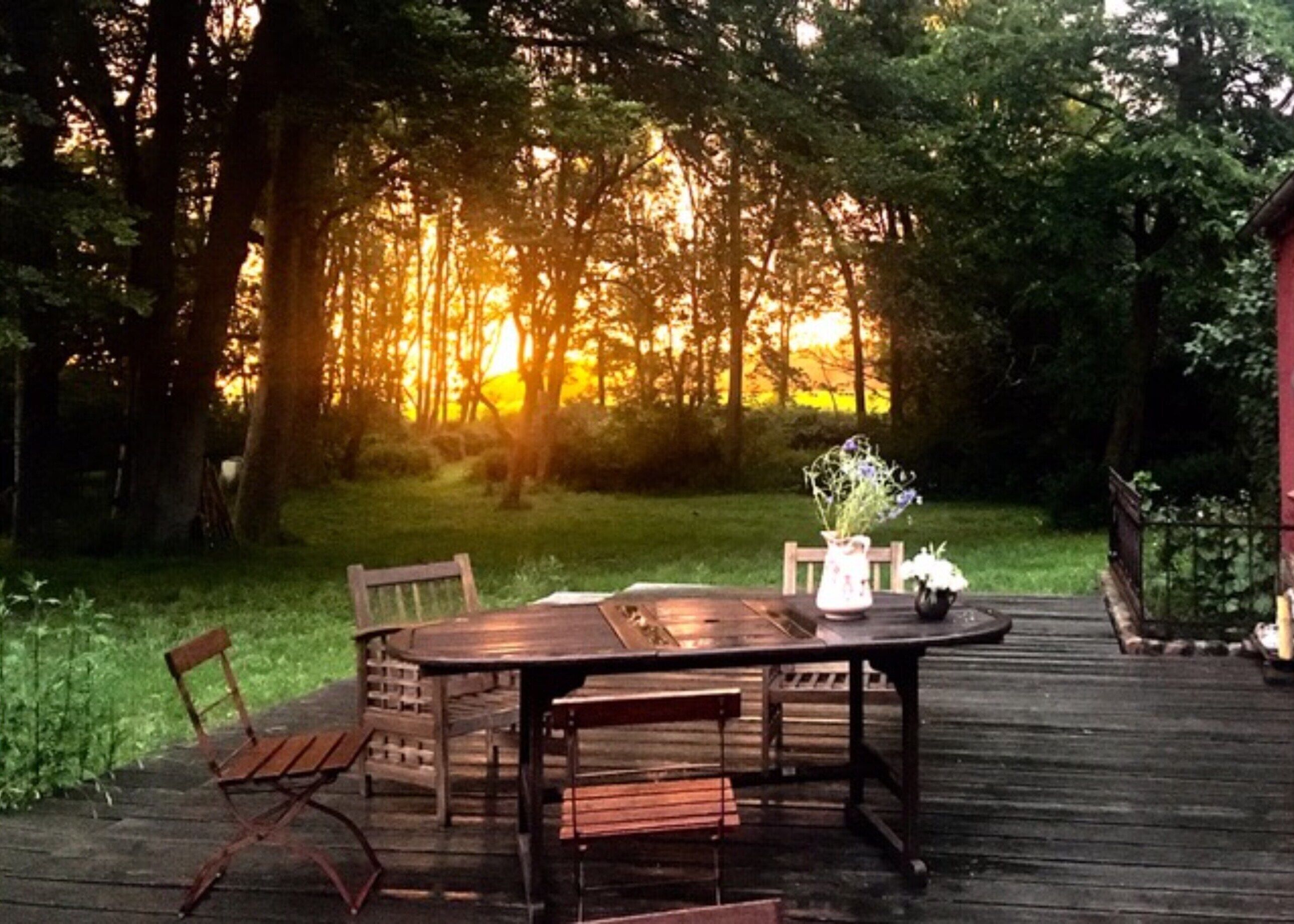 Een houten terras met een ronde tafel en stoelen, omgeven door een weelderig groonlandschap en bomen.