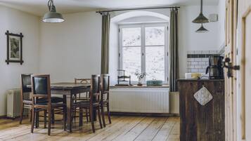 A dining area with a wooden table and chairs, a window with curtains, and a kitchen with a coffee maker.