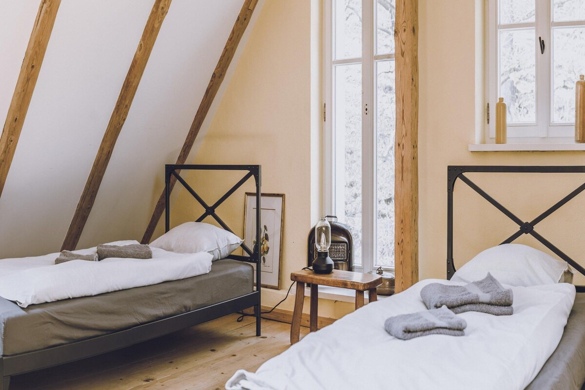 Une chambre confortable située dans les combles, avec deux lits, des poutres apparentes, une fenêtre donnant sur un beau paysage et une petite table avec une lampe.