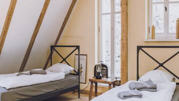 A cozy attic bedroom with two beds, wooden beams, a window with a view, and a small table with a lamp.