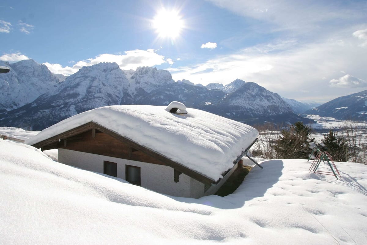 A snow-covered mountain cabin with a wooden exterior and a sloped roof, surrounded by snow-covered mountains and a clear sky.