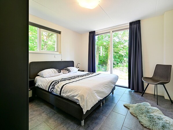 A bedroom with a bed, a chair, and a view of greenery through the windows.