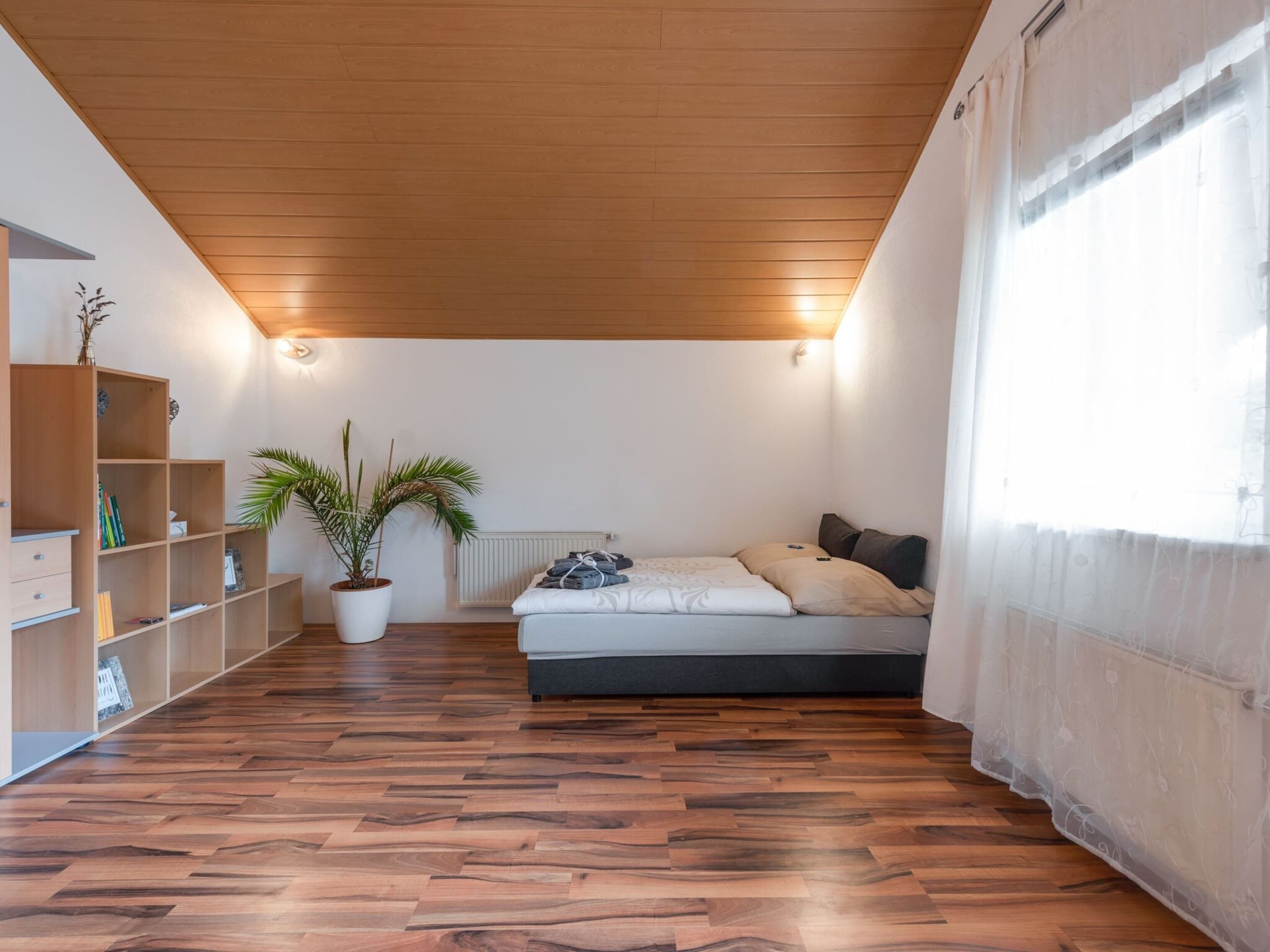 A room with a wooden ceiling, a bed with pillows, a radiator, a bookshelf, and a potted plant.