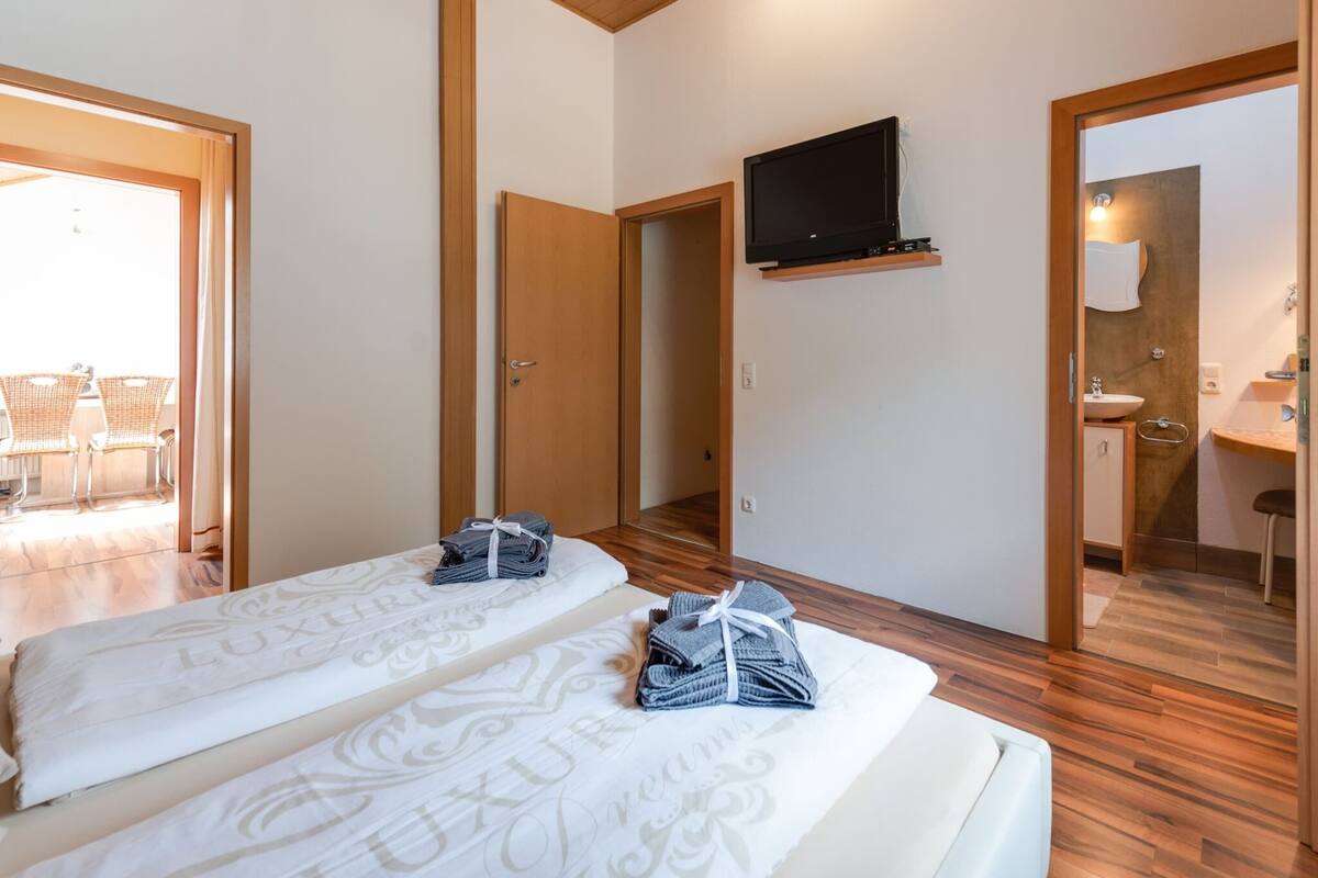 A hotel room with two beds, a TV mounted on the wall, a bathroom with a sink and mirror, and a balcony with chairs.