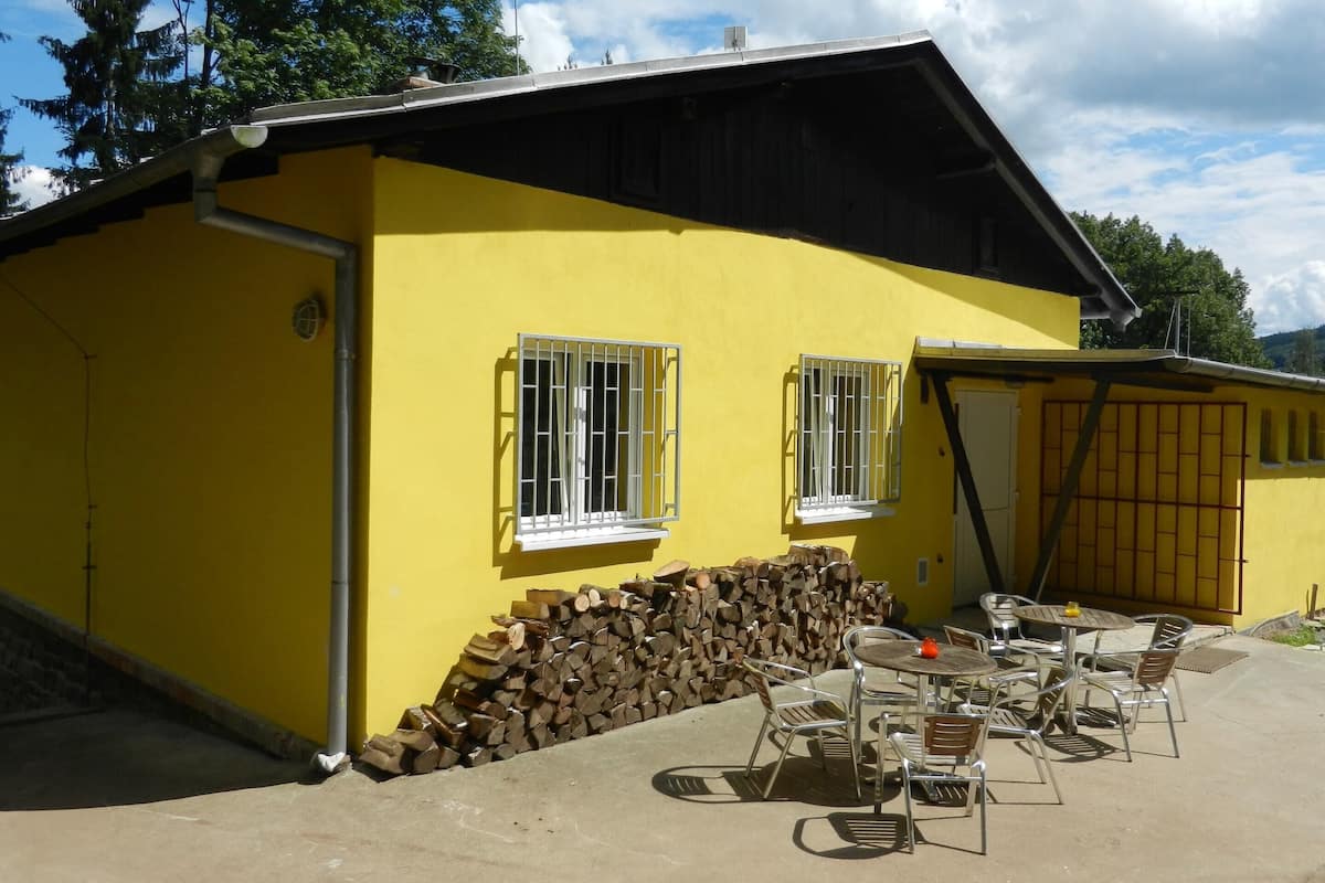 Un bâtiment jaune doté d’un espace extérieur abrité avec des tables et des chaises, et où est entreposée une pile de bois de chauffage.