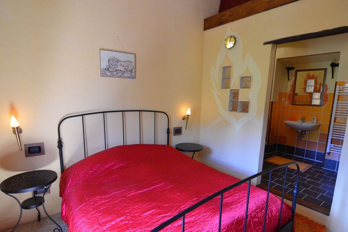A bedroom with a metal bed frame, a red bedspread, a small bedside table, a wall-mounted lamp, and a bathroom visible through an open door.