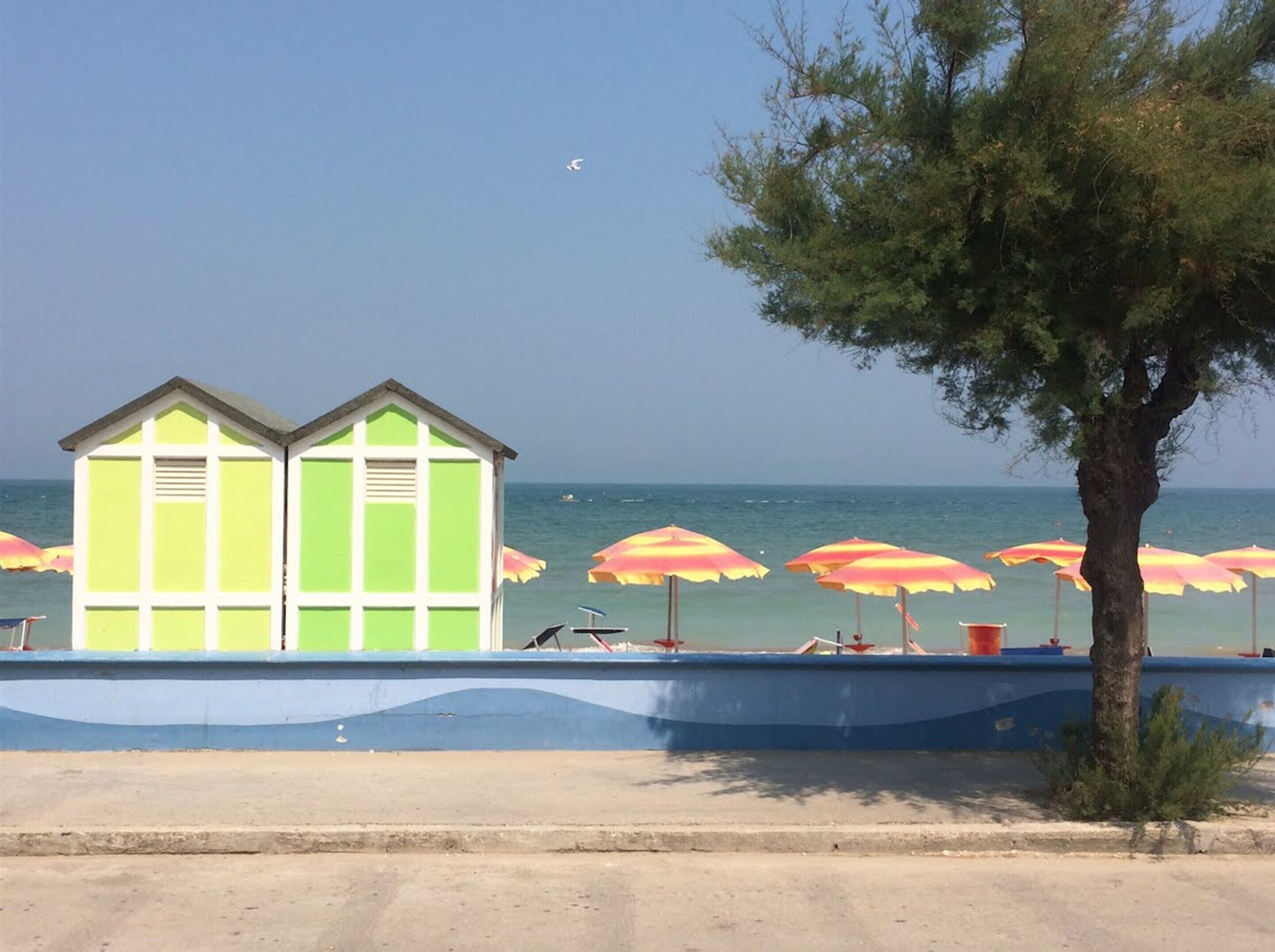 A beach with two green beach huts, umbrellas, and a tree.