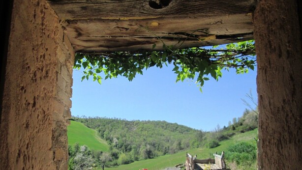Un arco rustico in pietra incornicia una vista di un sentiero sterrato che conduce a una struttura in legno con una panchina e piante in vaso.