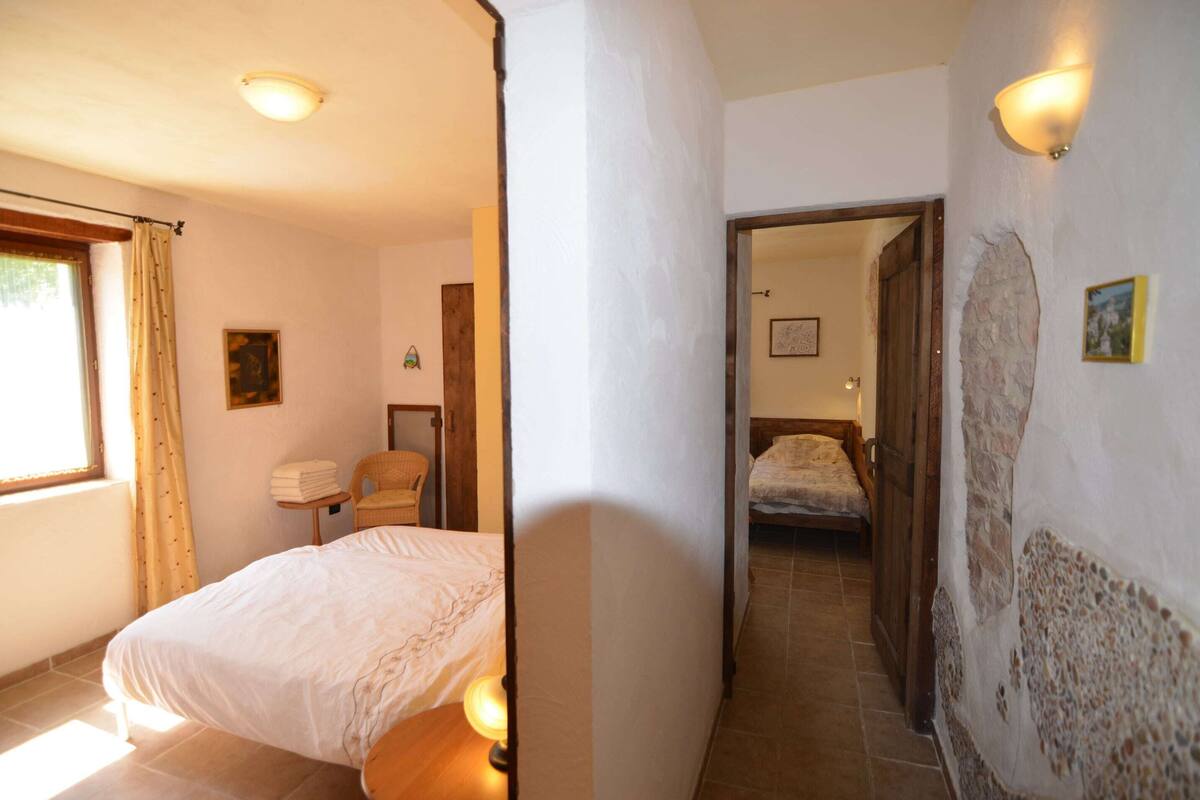 A bedroom with a bed, a chair, a small table, and a nightstand. There is a door leading to another room.