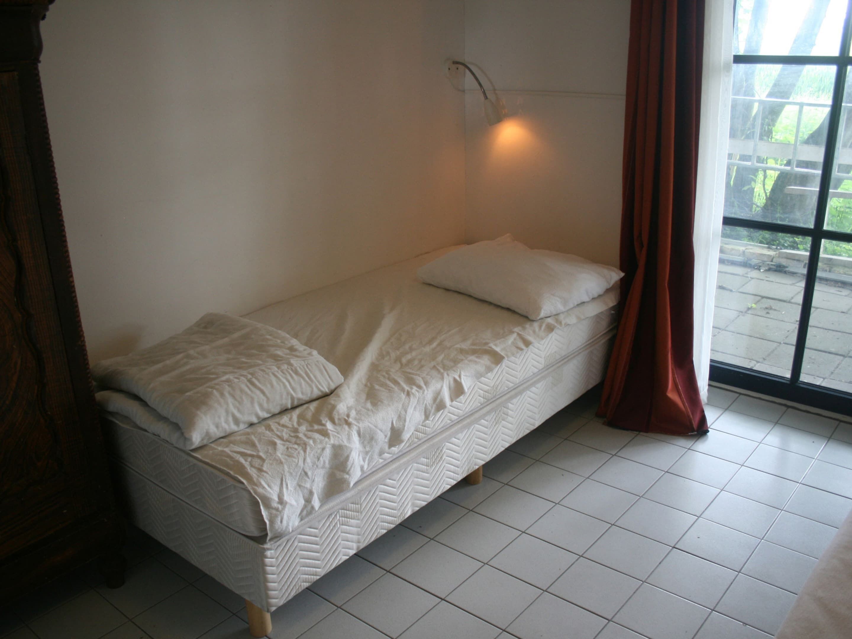 A single bed with a white mattress and pillows, a wooden headboard, a wall-mounted light fixture, and a window with a view of greenery outside.