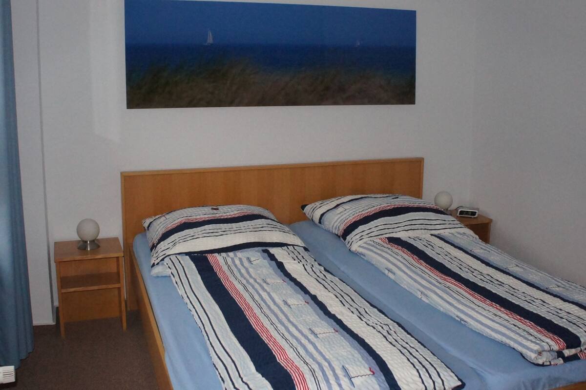 A bedroom with two single beds, a nightstand with a lamp, and a painting of a seascape on the wall.