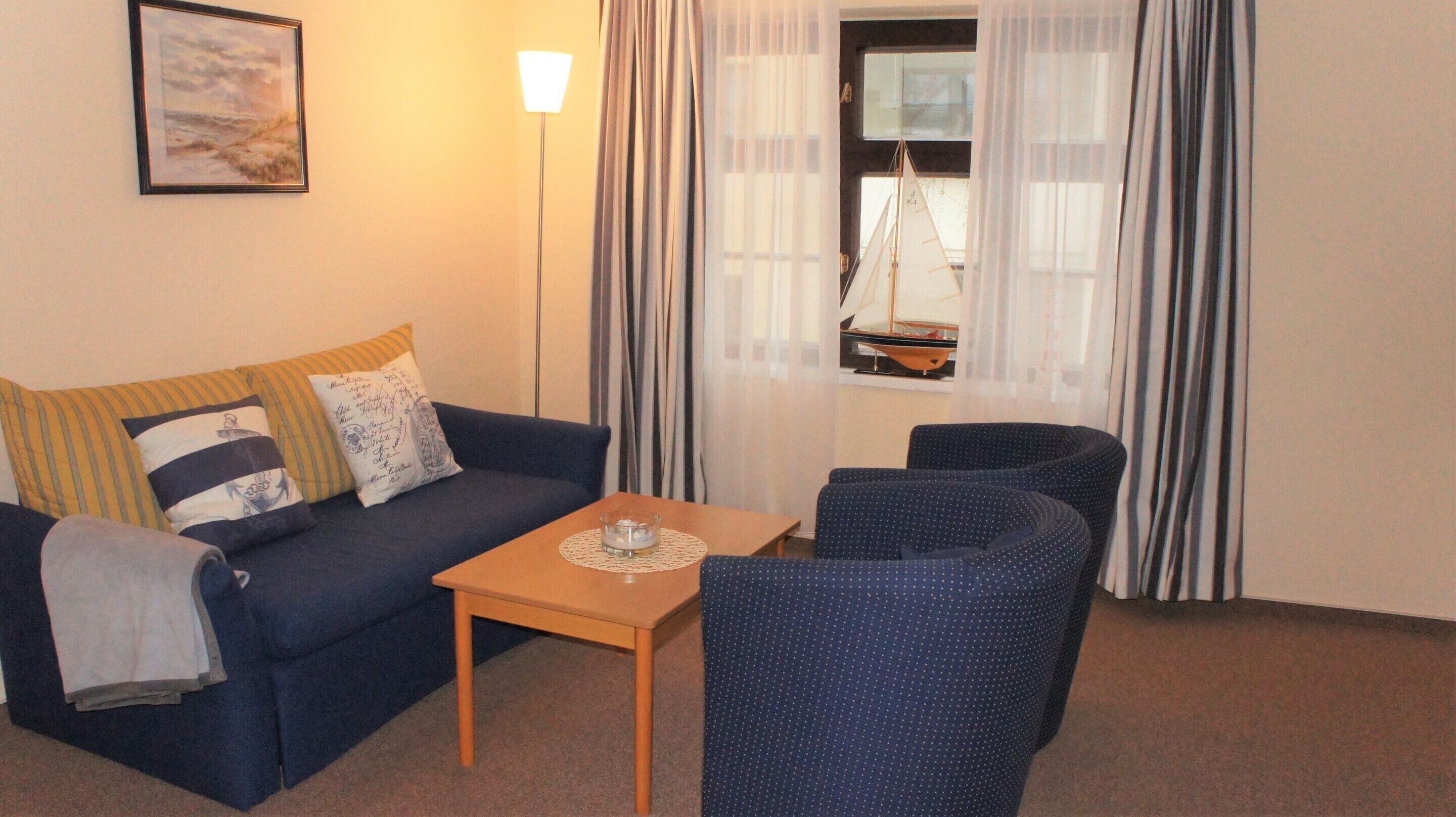 A living room with a sofa, a small table, and two armchairs. There is a window with curtains and a view of a sailboat outside.