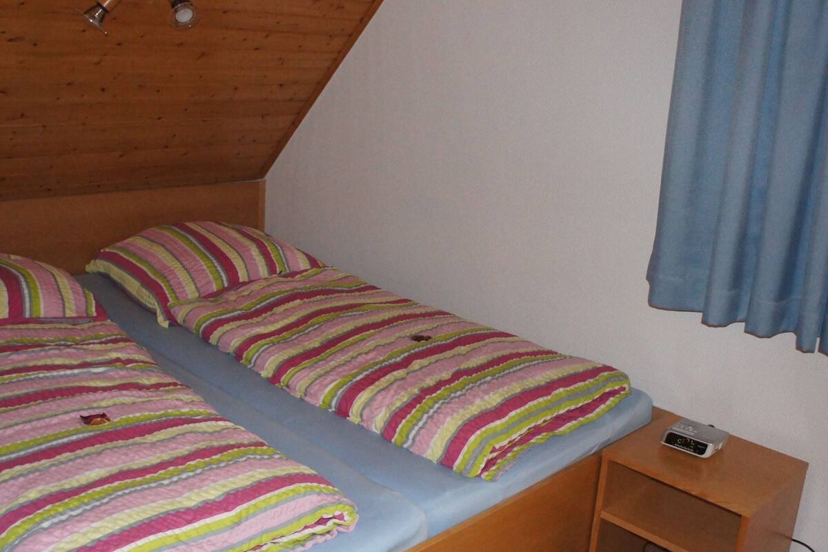 A small, single-bed room with a wooden ceiling, a blue curtain, and a bedside table with a clock.