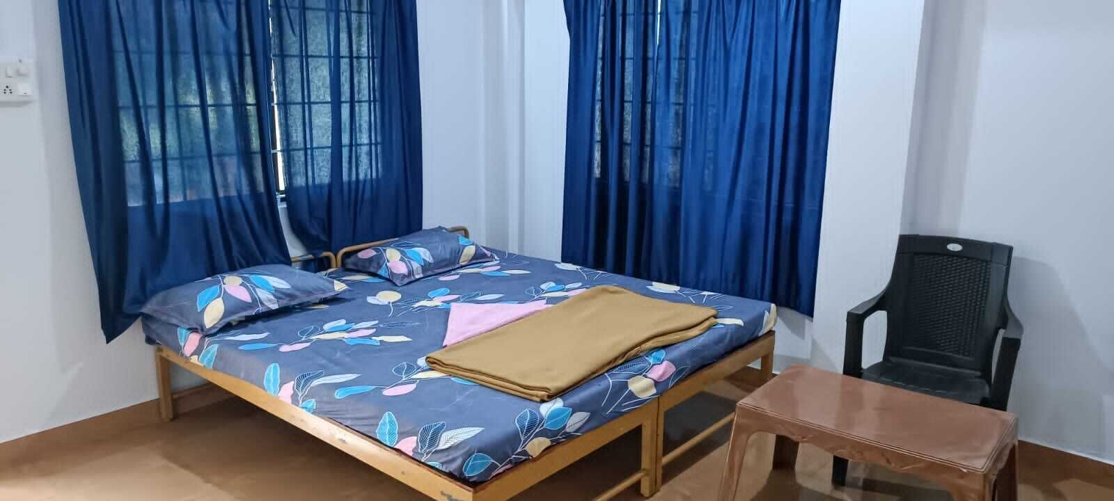 Una cama individual con colcha estampada, estructura de madera y manta marrón. Hay una mesita de madera al lado de la cama y una silla de plástico negro.
