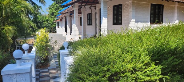 A pathway leading to a white building with a tiled roof and black shutters. - Swastha Wellness Centre (Kumta)