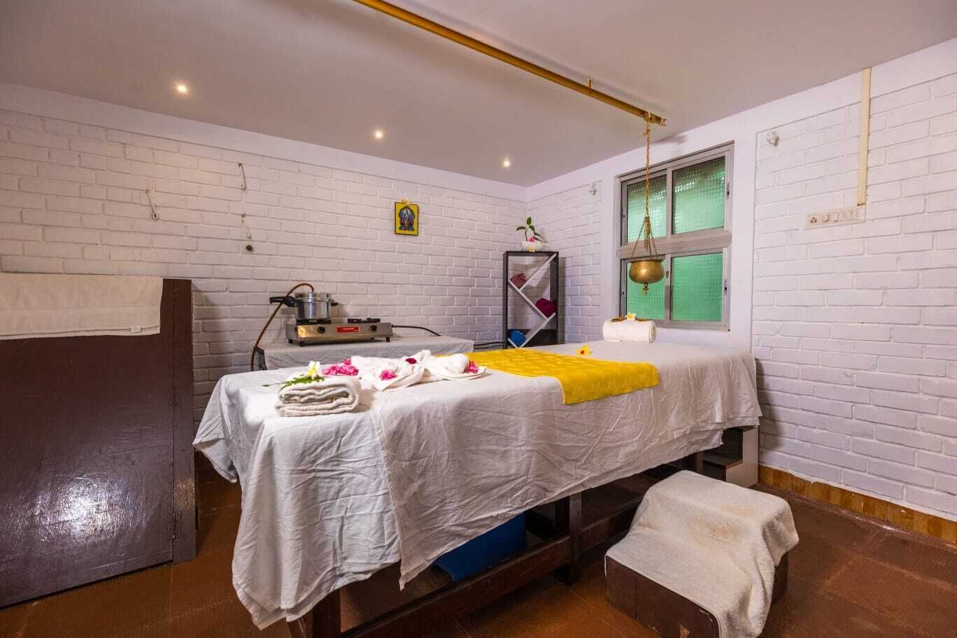 A single bed with a white bedspread, a yellow throw blanket, and towels. There is a small shelf with a lamp and a decorative item on the wall.