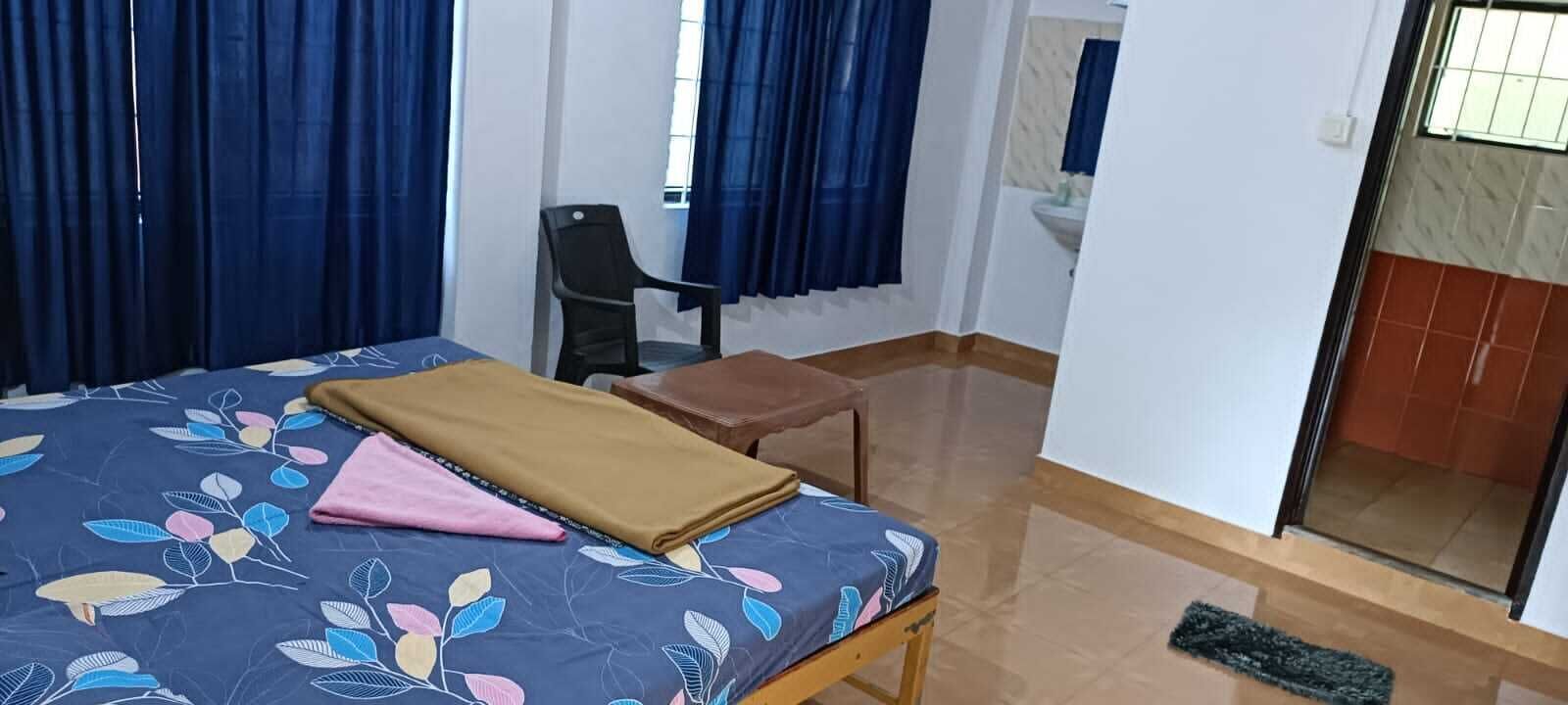Un dormitorio con cama, una silla, una mesita y un baño visible a través de una puerta abierta.