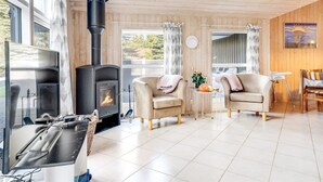 A cozy living room with a wood-burning stove, two leather sofas, a small table with flowers, and large windows offering a view of the outdoors.