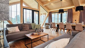 A spacious living area with a high ceiling, wooden beams, a sofa set, a dining table with chairs, and large windows offering a view of the outdoors. - Majestic Chalet With Pool and Sauna (Haus)