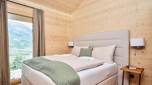 A neatly made bed with a green throw, a bedside table with a lamp, and a view of the outdoors through a large window. - Majestic Chalet With Pool and Sauna (Haus)