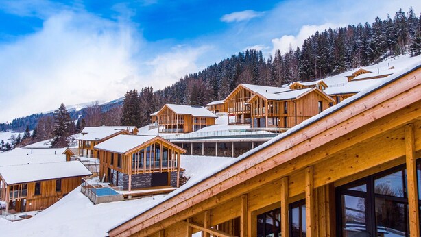 A cluster of wooden cabins with snow-covered roofs, set against a backdrop of a clear blue sky and a forested mountain. - Majestic Chalet With Pool and Sauna (Haus)