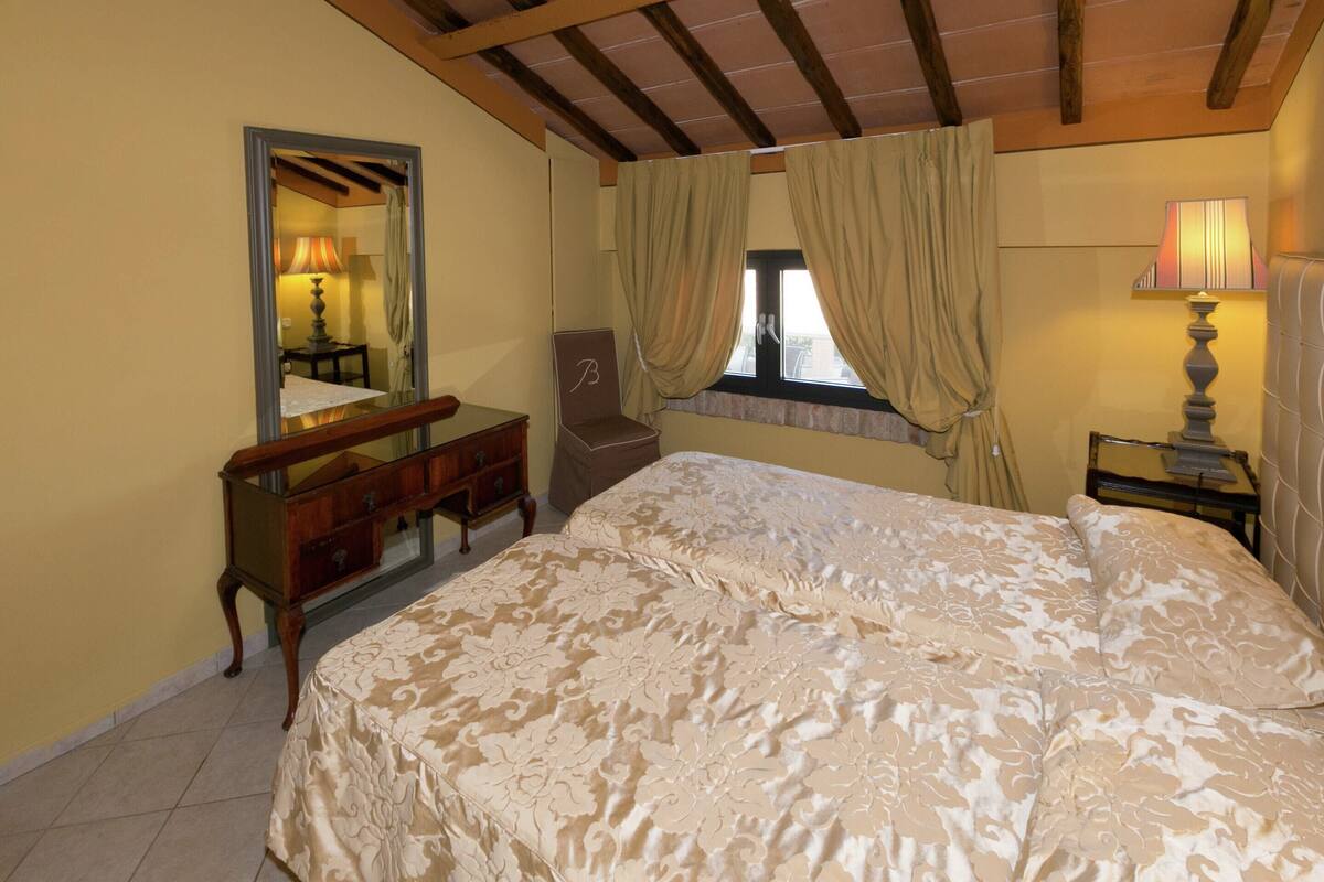A bedroom with a large bed, a wooden dresser, a chair, a window with curtains, and a lamp.