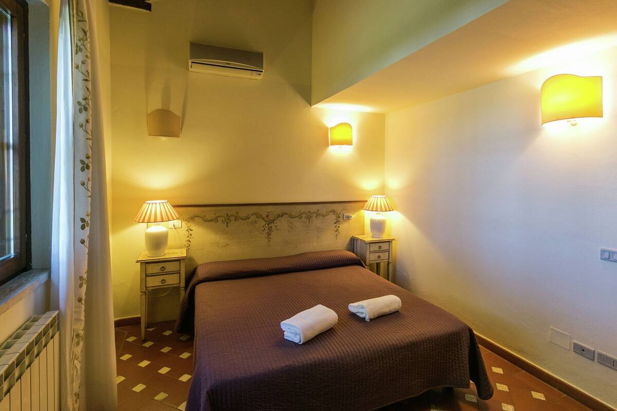 A hotel room with a bed, two bedside lamps, a window with curtains, an air conditioning unit, and a wall-mounted light fixture.