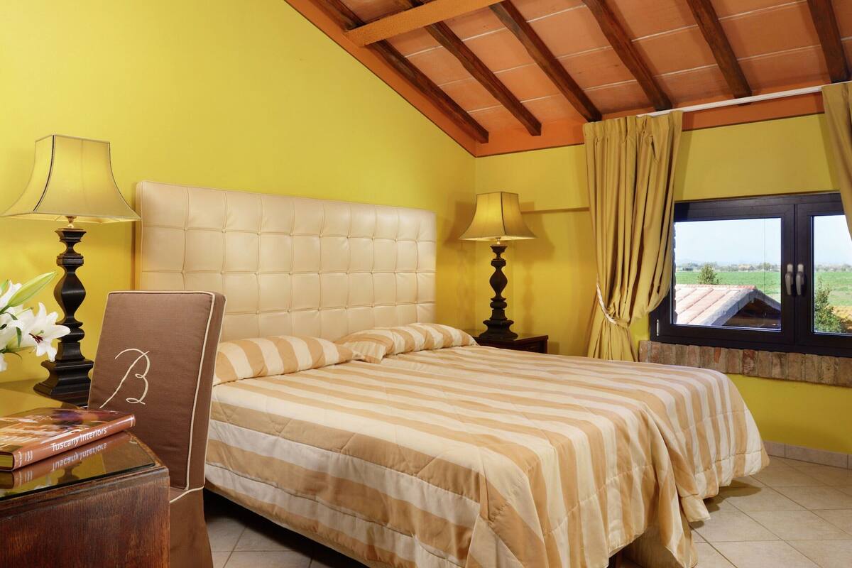 A bedroom with a large bed, two bedside lamps, a nightstand, a window with curtains, and a view of the outside.