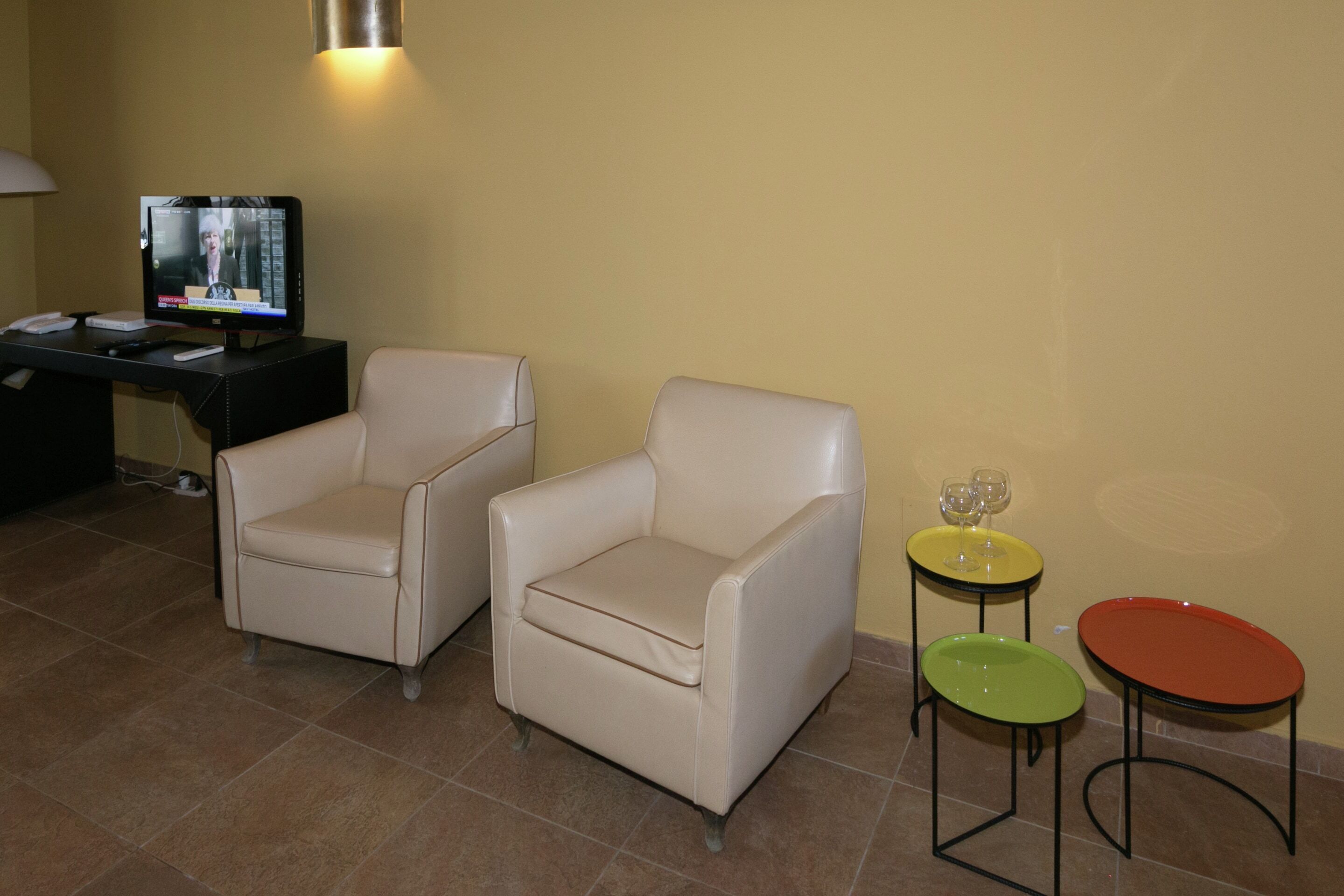 A room with two beige armchairs, a black desk with a television, and two small tables with wine glasses.