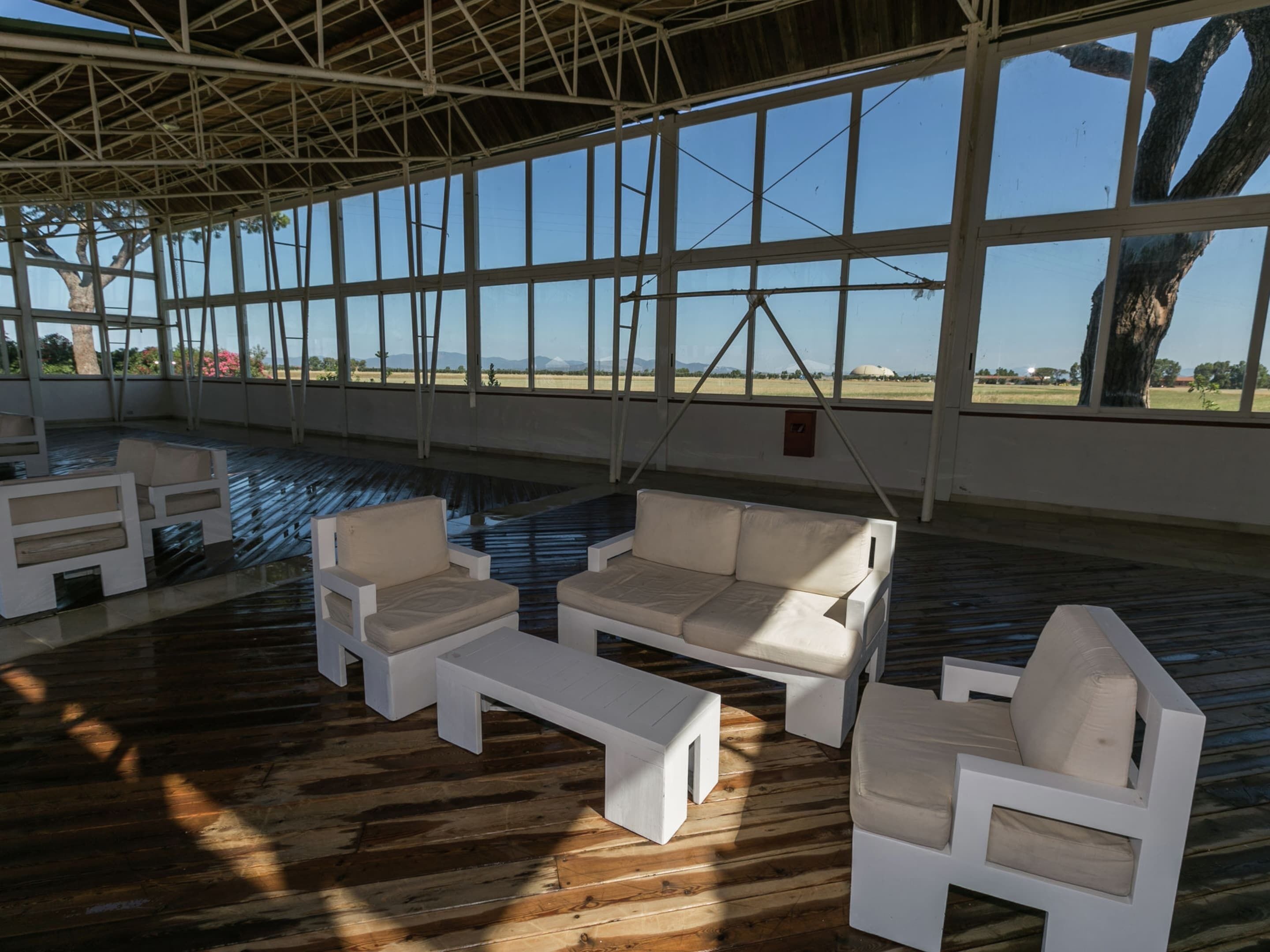 A spacious indoor area with large windows, wooden flooring, and white furniture including sofas and a coffee table.