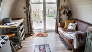 A cozy cabin interior with a wooden interior, a brown leather sofa, a small desk with a chair, and a sliding glass door leading to an outdoor area. - Rutland Rural Retreats - Woodpecker pod
 (Esher)