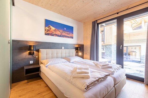 A bedroom with a large bed, a headboard, two bedside tables, a wall-mounted lamp, and a painting of a mountain landscape.