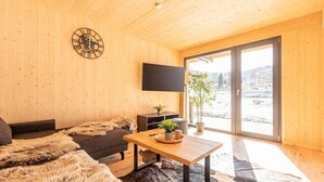Ein gemütliches Blockhaus-Interieur mit Holzbalkendecke, einer Wanduhr, einem Flachbildfernseher, einer Couch, einem Couchtisch und einem großen Fenster mit Blick auf eine verschneite Landschaft.