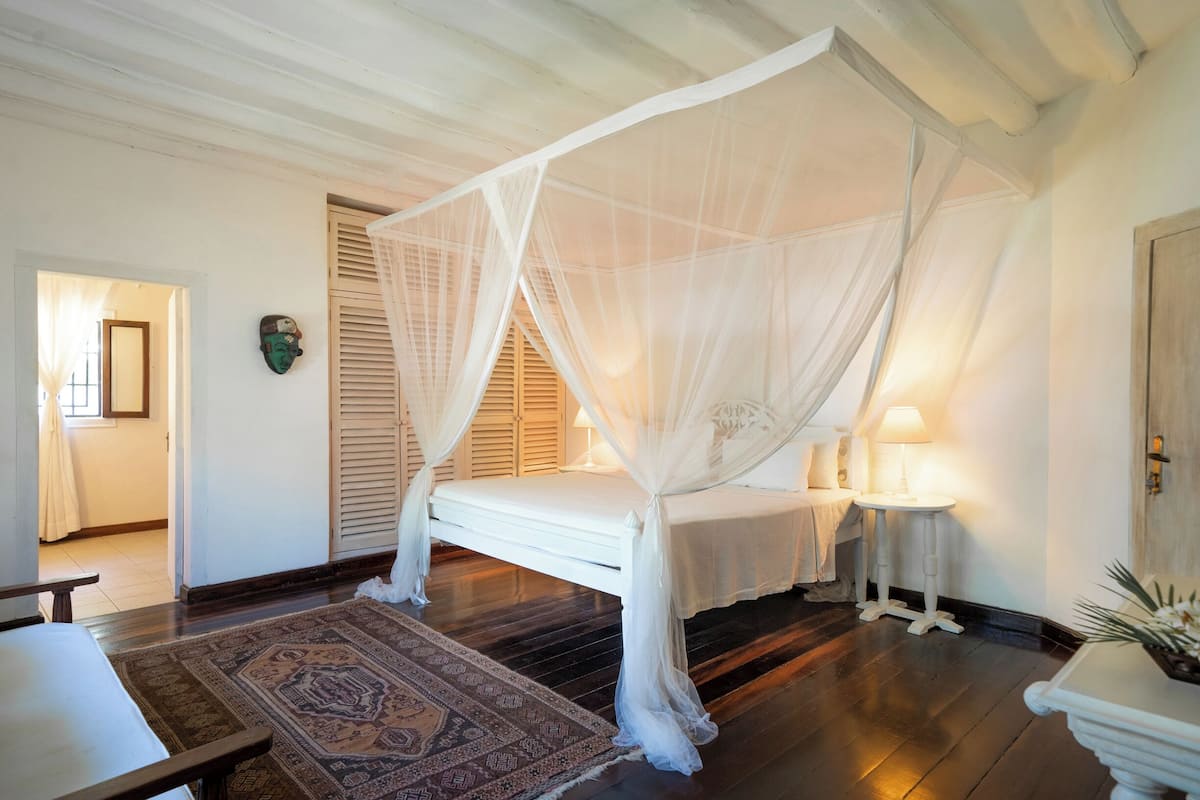 A room with a four-poster bed, a small bedside table, a nightstand, a patterned rug, and a door with a shutter.