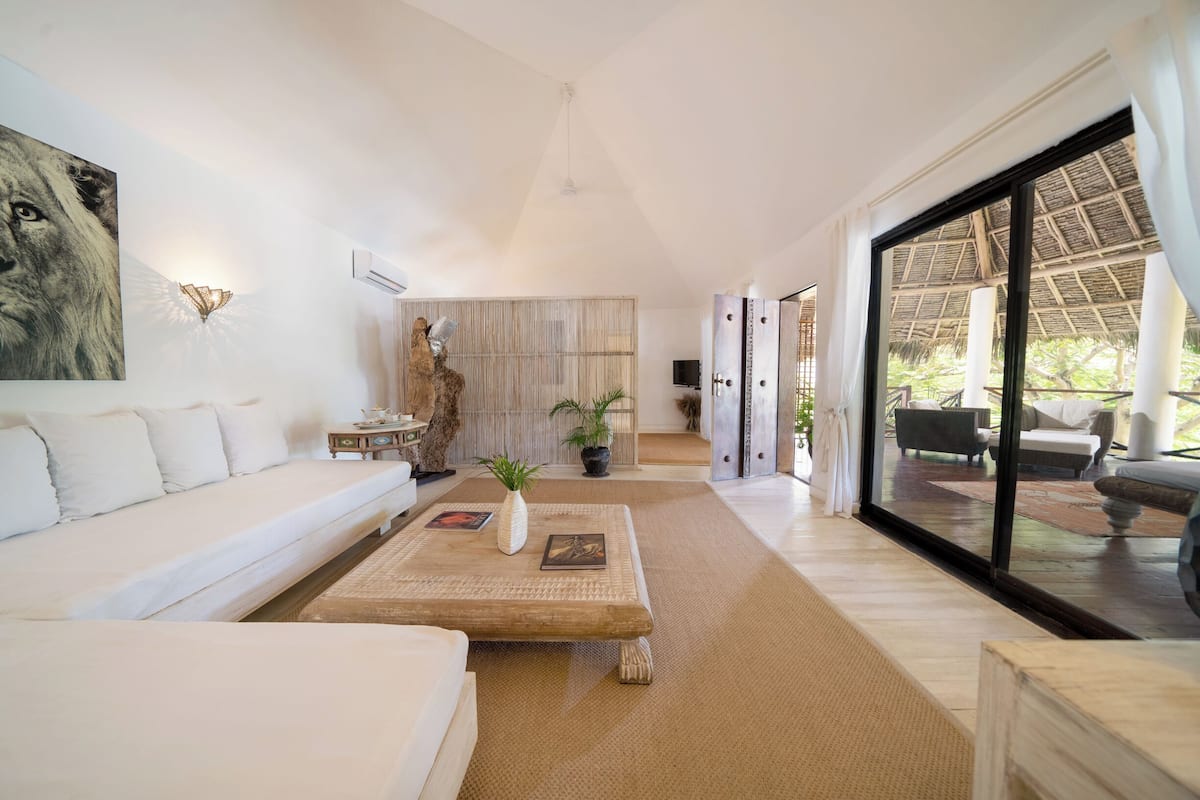 A spacious living room with a white sofa, a wooden coffee table with decorative items, a large window with a view of the outdoors, and a modern ceiling design.