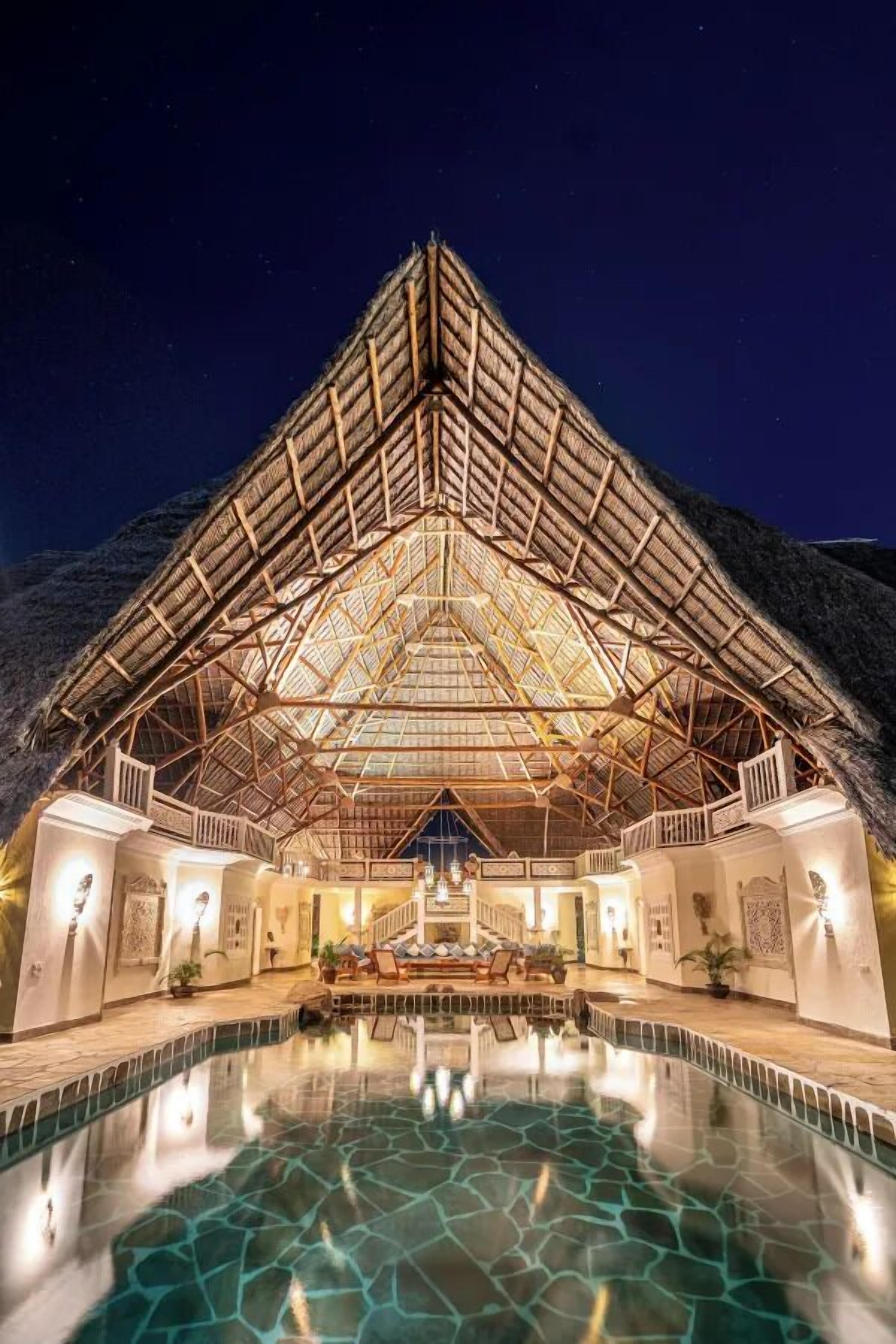 A large, open-air structure with a glass ceiling, a swimming pool in front, and a mountainous backdrop.