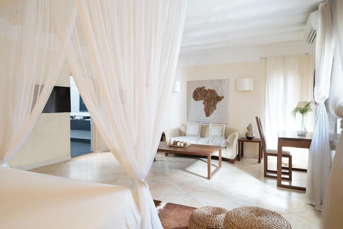 A room with a bed, a sofa, a coffee table, and a dining table. There is a large wall art piece and a plant on the dining table.