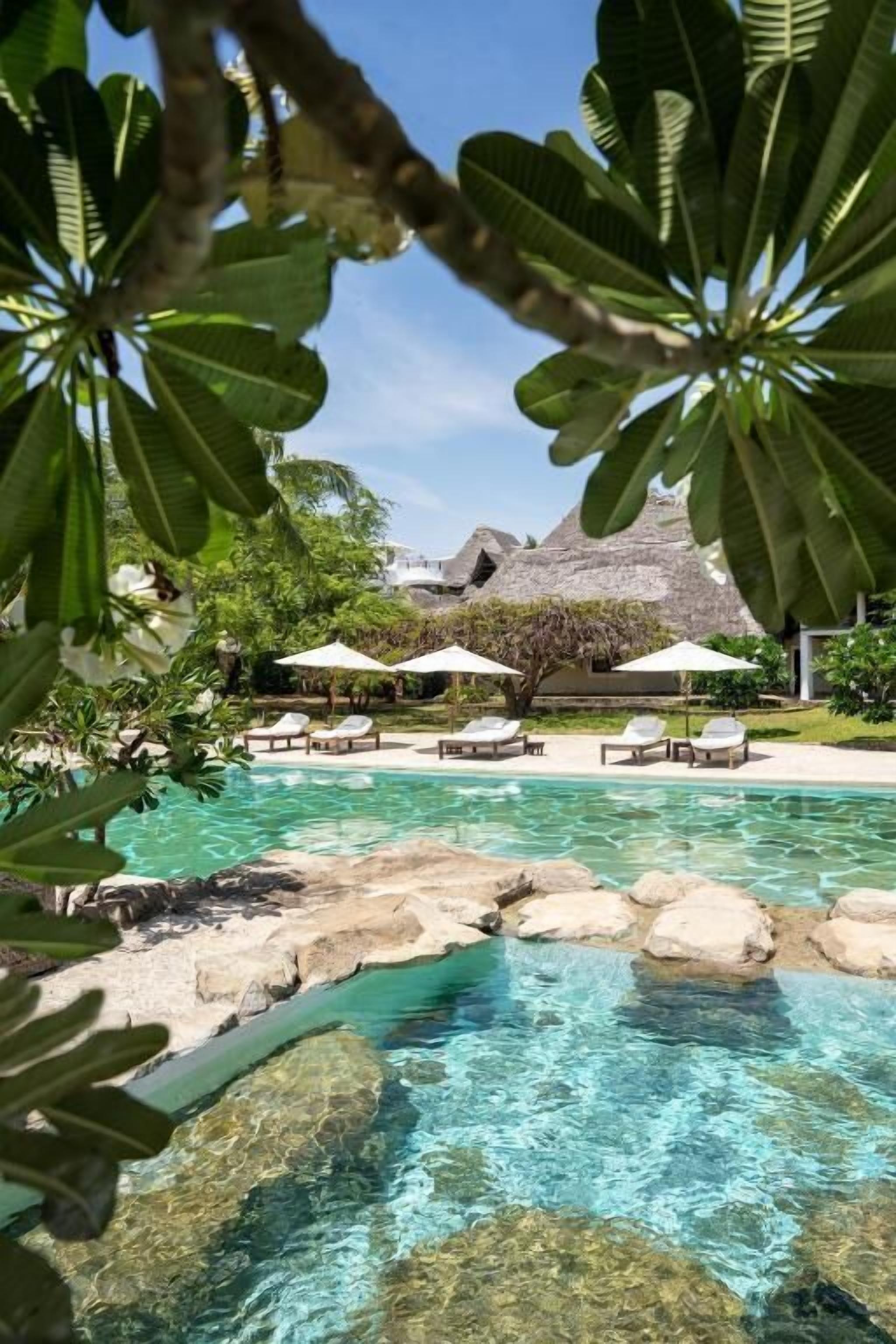 Une piscine aux eaux cristallines, bordée de rochers, avec des chaises longues, des parasols et, au fond, une structure au toit de chaume.