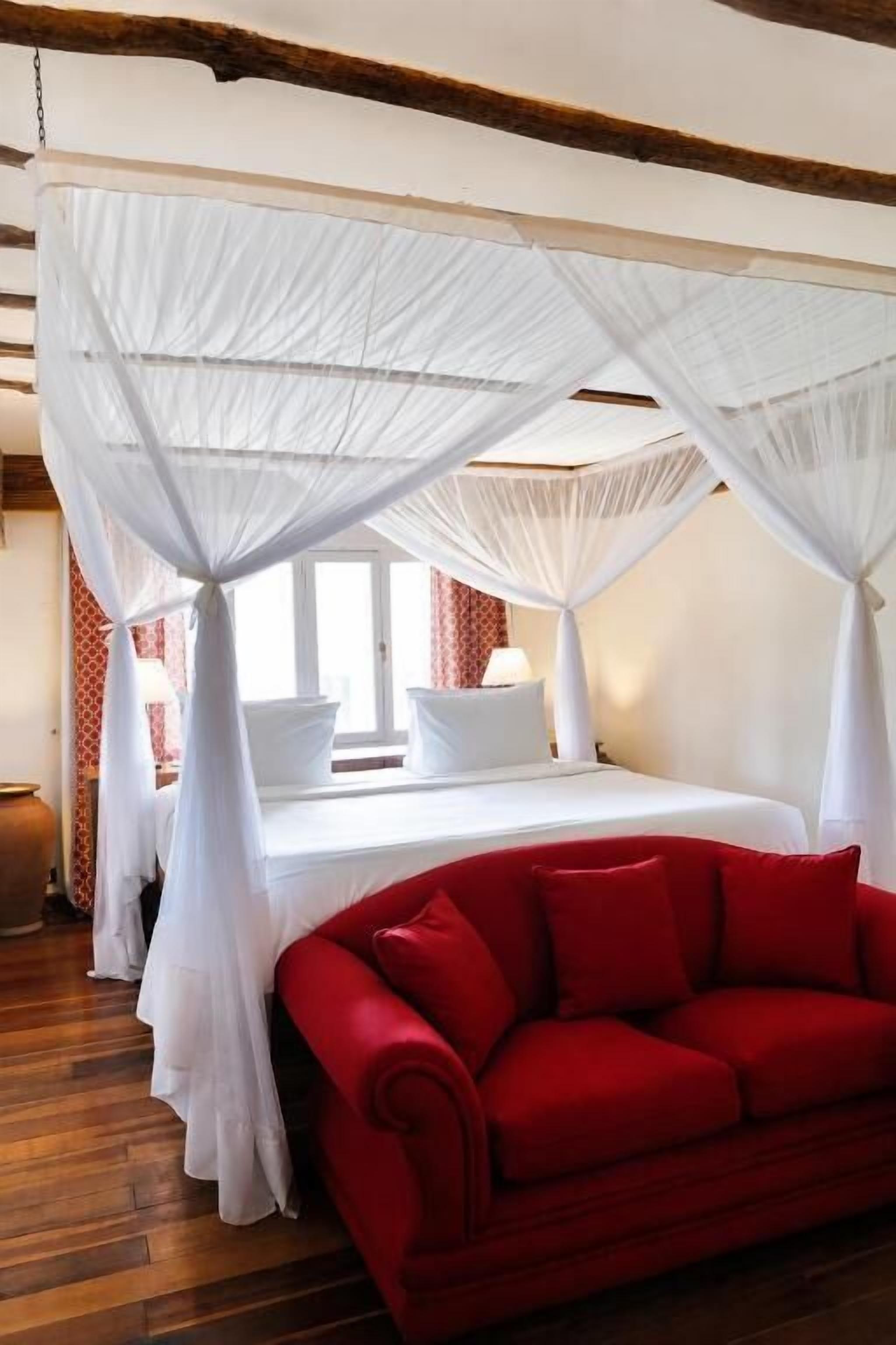 A room with a bed under a white canopy, a red sofa, and a window with curtains.