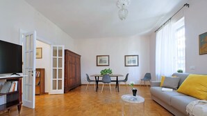 A spacious living room with a wooden parquet floor, a grey sofa with yellow cushions, a dining table with chairs, a television, and a bookshelf. - 4bnb - Corridoni Apartment (Rome)