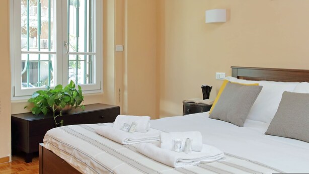 A neatly made bed with white linens and towels, a bedside table with a plant, and a window with a view of buildings outside. - 4bnb - Corridoni Apartment (Rome)