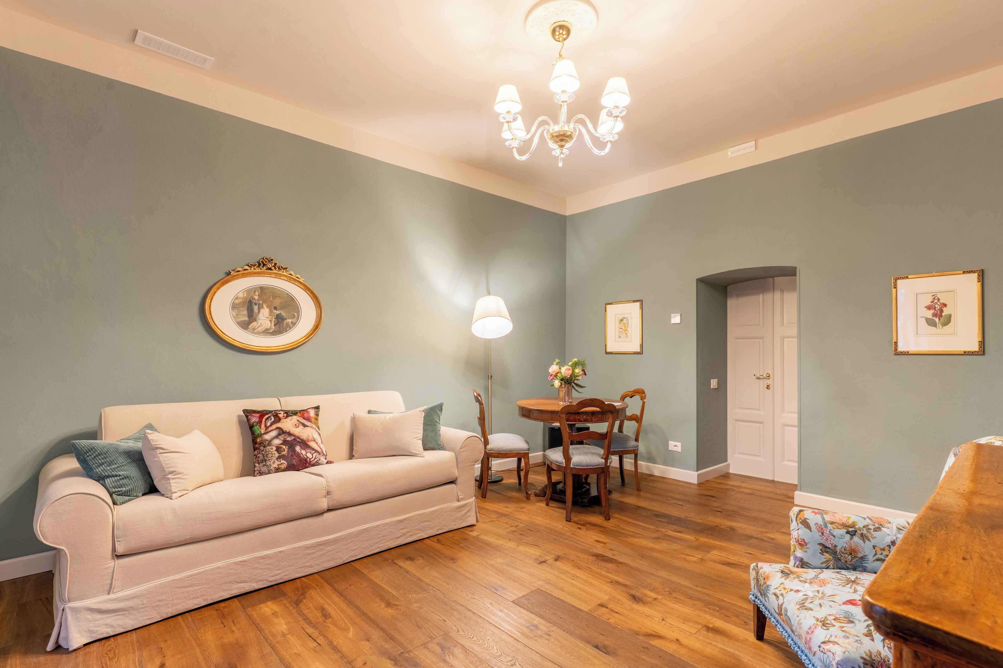 Un salon avec un canapé beige, une table ronde en bois et des chaises. Un lustre est suspendu au-dessus de la table et un tableau encadré orne le mur.