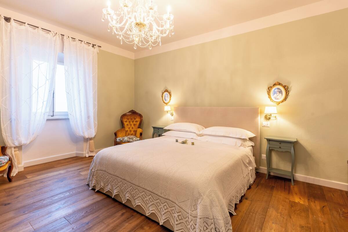 Ein Schlafzimmer mit einem großen Bett, zwei Nachttischen mit Lampen, einer Chandelier und einem Sessel.