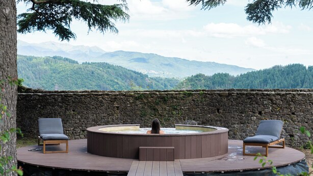 Un bain à remous sur une terrasse en bois, avec deux chaises, entouré d’un mur de pierre et offrant une vue sur les montagnes.