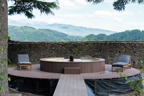Un bain à remous sur une terrasse en bois, avec deux chaises, entouré d’un mur de pierre et offrant une vue sur les montagnes.