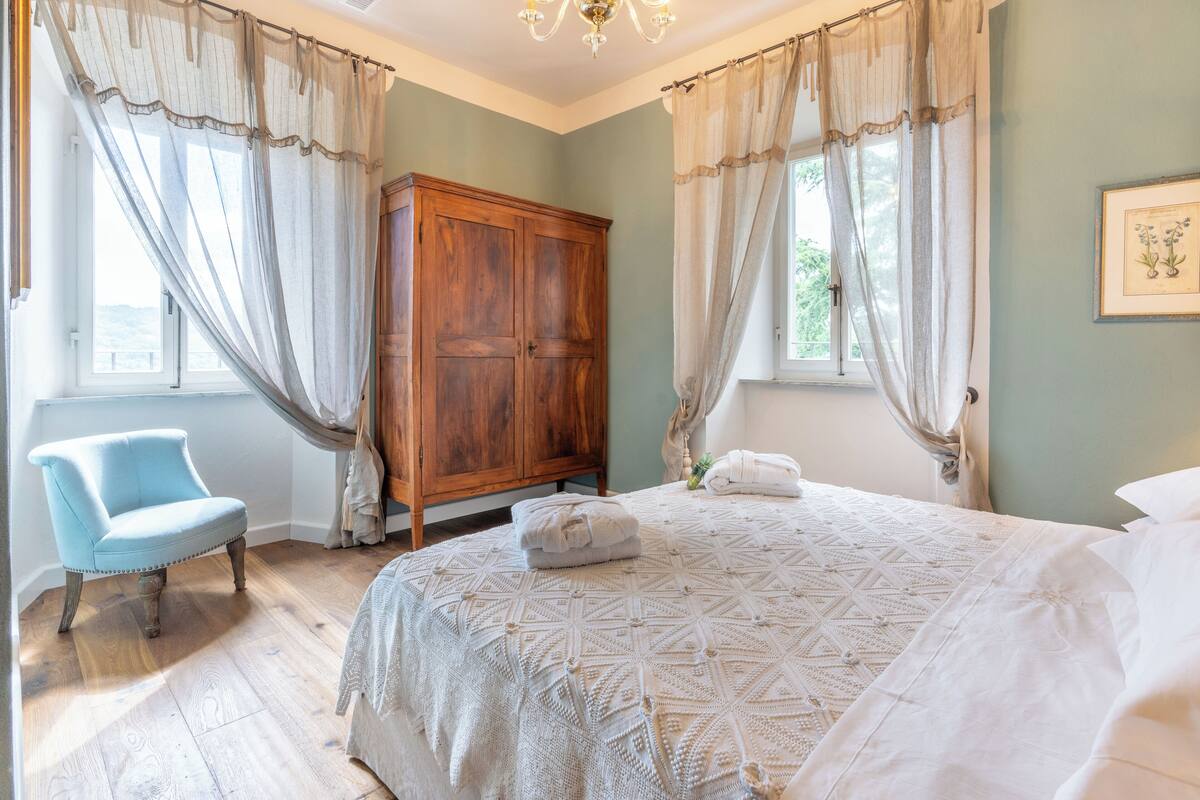 Ein Schlafzimmer mit einem großen Bett, einem Holzschrank, einem blauen Sessel und einem Fenster mit Vorhängen.
