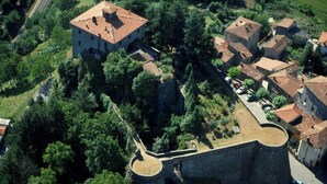 Une vue aérienne d’une ville historique, dominée par une imposante fortification en pierre, entourée d’une végétation luxuriante et de bâtiments traditionnels aux toits couverts de tuiles rouges.