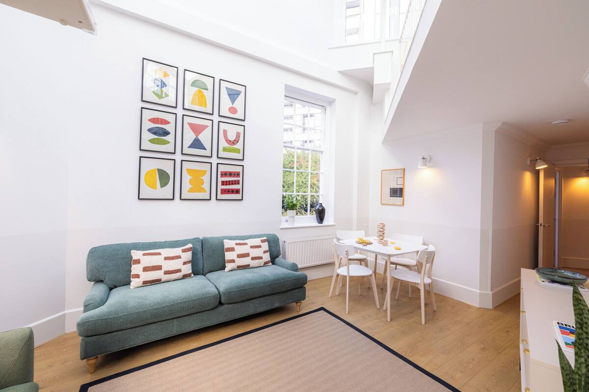 A modern living room with a teal sofa, a dining table with chairs, and a large window.